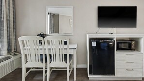 Ett rum med ett skrivbord, två vita stolar, ett svart kylskåp, en mikrovågsugn, en platt-TV och en spegel.