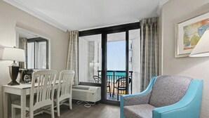 A room with a dining table, chairs, a blue armchair, a large window with curtains, and a framed picture on the wall. - Ocean View King Room + Beach Access + Lazy River (Myrtle Beach)