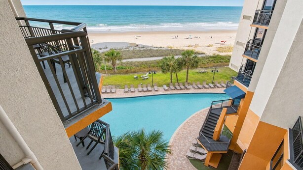 A pool area with a view of the beach and ocean, a small lawn with lounge chairs, and a building with balconies. - Ocean View King Room + Beach Access + Lazy River (Myrtle Beach)
