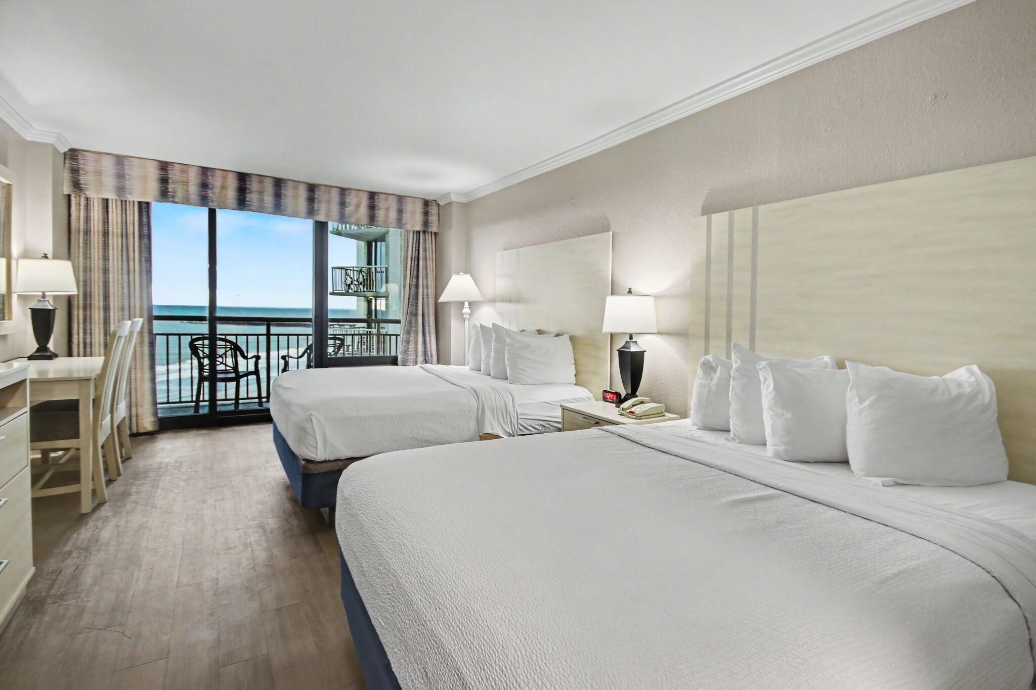 A hotel room with two beds, a desk, a chair, a phone, and a balcony with a view of the sea.