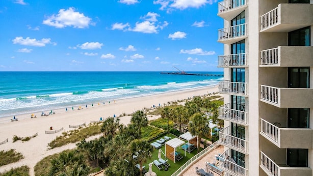 A beachfront hotel with balconies, a sandy beach, and a pier in the background. - Oceanfront Studio w/ Two Queens + Beach & Pools (Myrtle Beach)