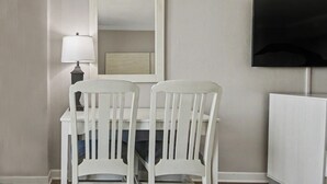 Una habitación con un escritorio blanco, dos sillas blancas, una lámpara, un espejo y un televisor en la pared.