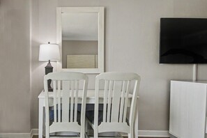 Ein Zimmer mit einem weißen Schreibtisch, zwei weißen Stühlen, einer Lampe, einem Spiegel und einem Fernseher an der Wand.