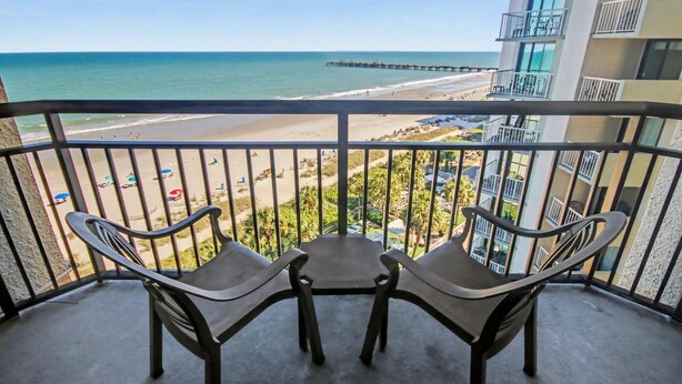 Un balcón con dos sillas y una mesa, con vista a una playa con muelle, un edificio con balcones y un cielo despejado.
