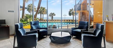 A modern hotel lobby with a pool view, two blue armchairs, a hexagonal coffee table, and a business center sign.