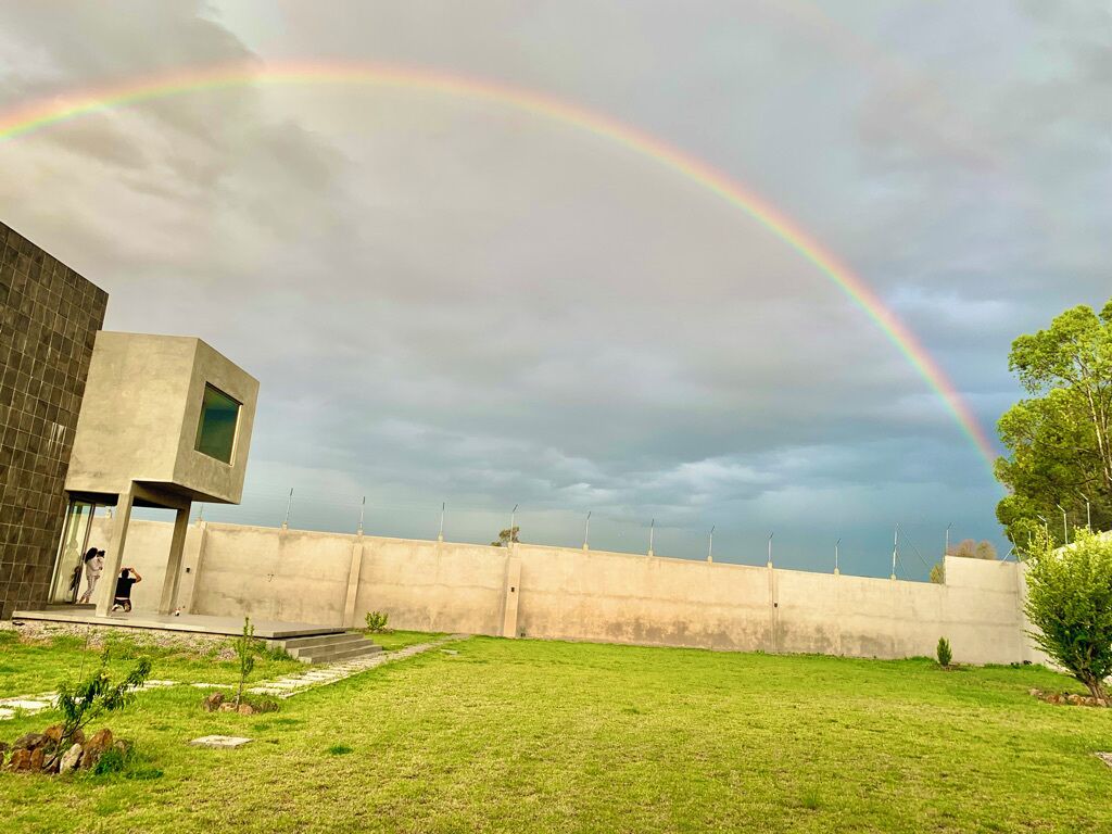 A modern building with a flat roof, a concrete wall, and a grassy area with a rainbow in the sky.