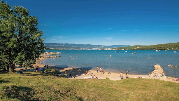 Een strand met mensen die zonnebaden en zwemmen, een heldere blauwe lucht en boten op het water.