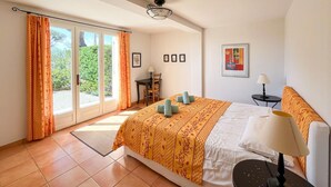 Ein Schlafzimmer mit Bett, Schreibtisch, Stuhl, Lampe und Blick auf Grün durch das Fenster.