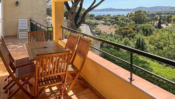 Ein Balkon mit Tisch und Stühlen, einem Geländer und Blick auf eine grüne Landschaft mit Bäumen und einer entfernten Stadtansicht.