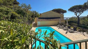 Una piscina con borde de baldosas, rodeada de vegetación y un edificio con techo de tejas.