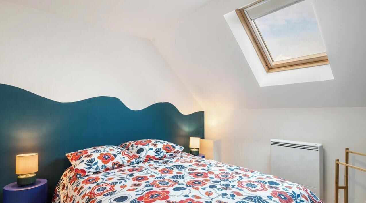 Quarto com cama, mesa de cabeceira, luminária, radiador e claraboia.