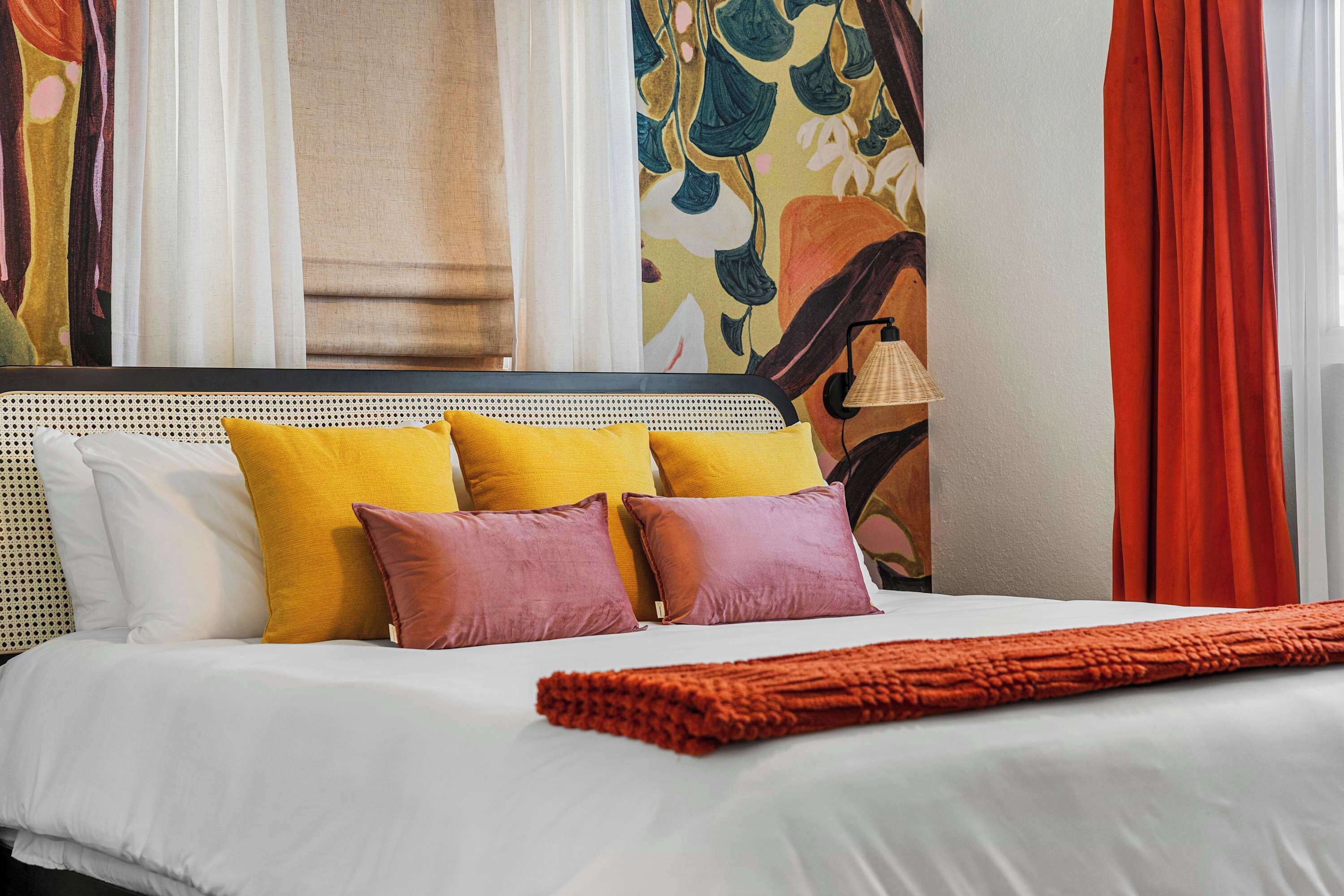 Un letto con biancheria bianca, numerosi cuscini giallo e viola, una coperta a maglia rossa e una parete con arazzo floreale.