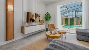 A modern living room with a flat-screen TV, a round wooden coffee table, a grey sofa, and a view of a swimming pool through large windows.
