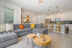 A modern living room with a grey sectional sofa, round wooden coffee table, and a kitchen area with white cabinets and a bar stool.