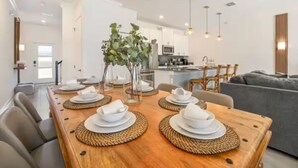 A modern dining area with a wooden table set for four, a vase with green foliage, and a kitchen in the background.