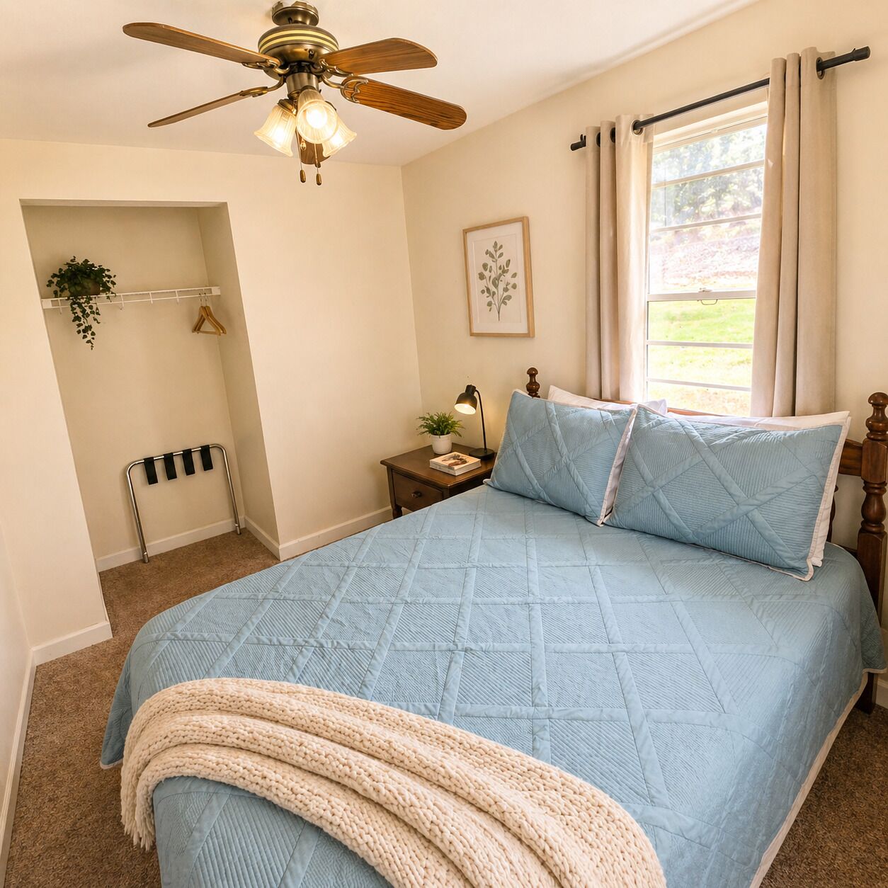 A bedroom with a bed, a ceiling fan, a bedside table with a lamp, a window with curtains, and a plant on the wall.