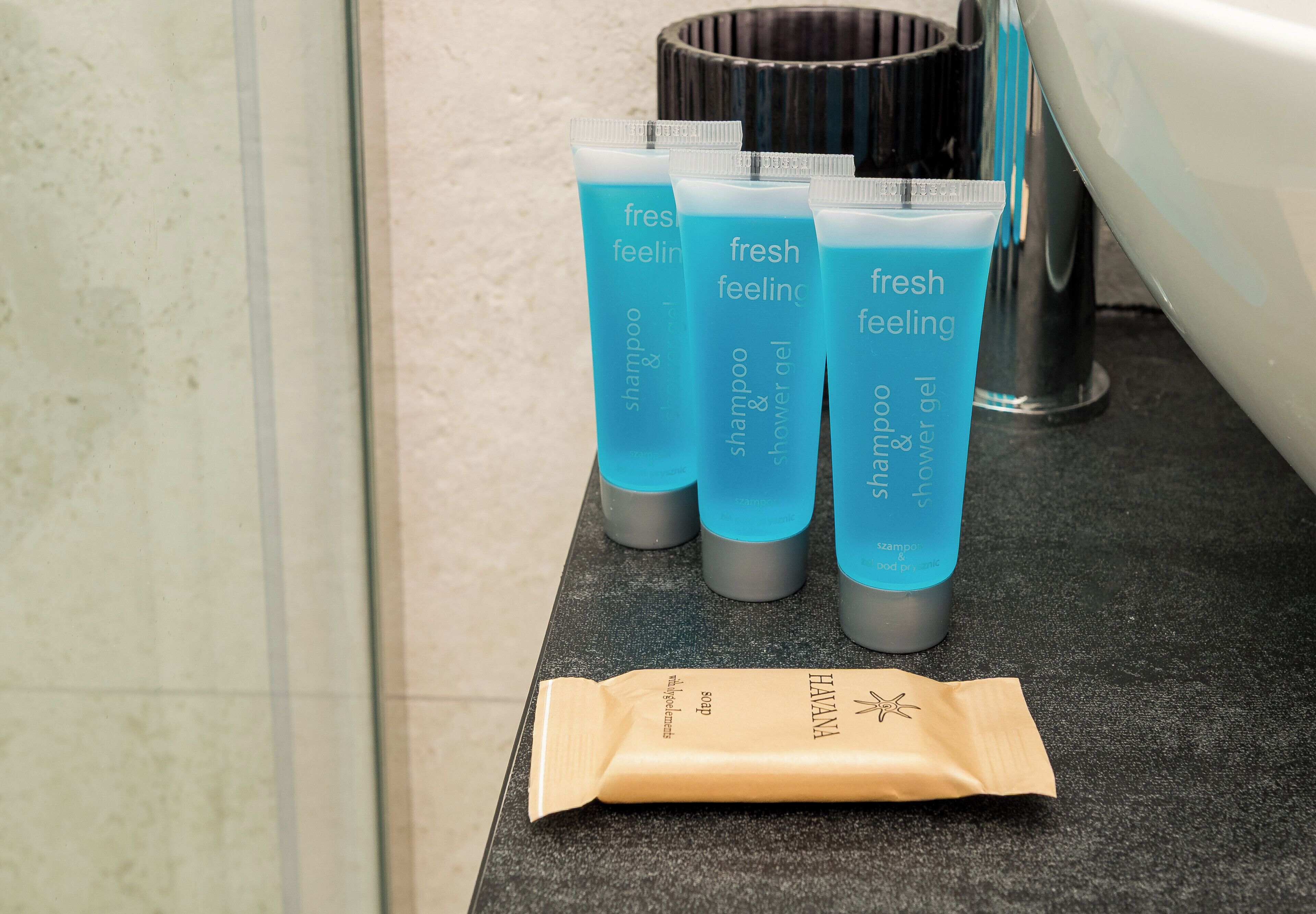 Uma bancada de banheiro com três frascos de shampoo e gel de banho 'fresh feeling', um recipiente de metal e um saco de papel marrom.