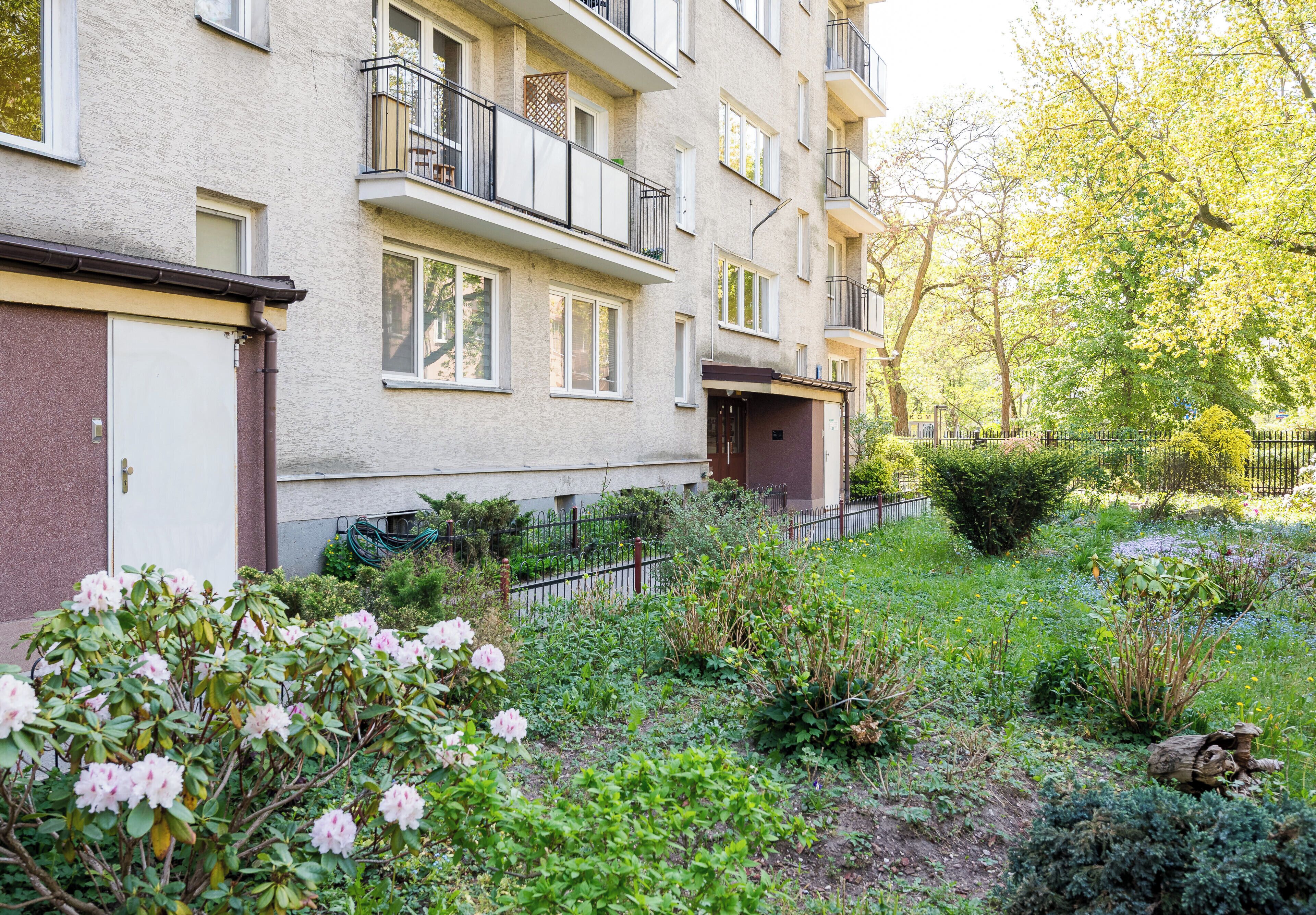 Un edifici residencial de diversos pisos amb balcons, un jardí amb diferents plantes i una tanca.