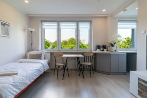 A compact room with a bed, a desk, two chairs, a small kitchenette, and large windows with a view of greenery. - Cichociemnych Chic Studio (Gliwice)
