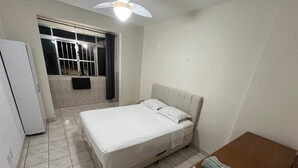 Un dormitorio con cama, una mesita de noche de madera, una ventana con cortinas, un ventilador de techo y un televisor montado en la pared.