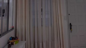 Una habitación con puerta blanca, una ventana con cortinas translúcidas, una lámpara en el techo y un estante con objetos.
