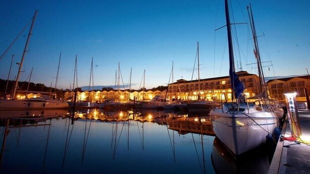 A marina with multiple boats docked, a building with warm lighting, and calm water reflecting the boats. - Lindner Hotel Boltenhagen (Boltenhagen)