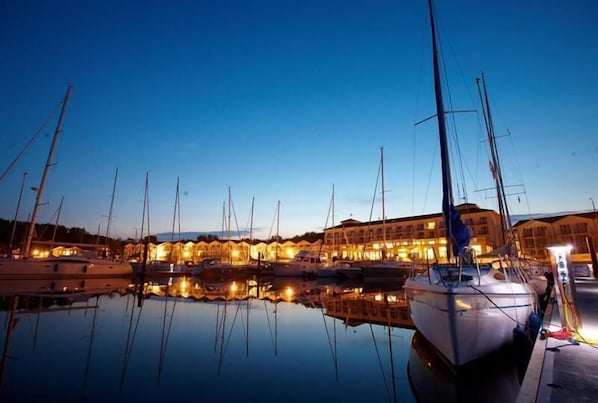 A marina with multiple boats docked, a building with warm lighting, and calm water reflecting the boats. - Lindner Hotel Boltenhagen (Boltenhagen)