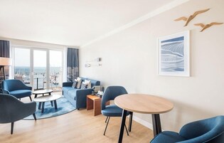 Una sala de estar moderna con sofá, sillas, una mesa redonda y vistas a la ciudad a través de amplios ventanales.