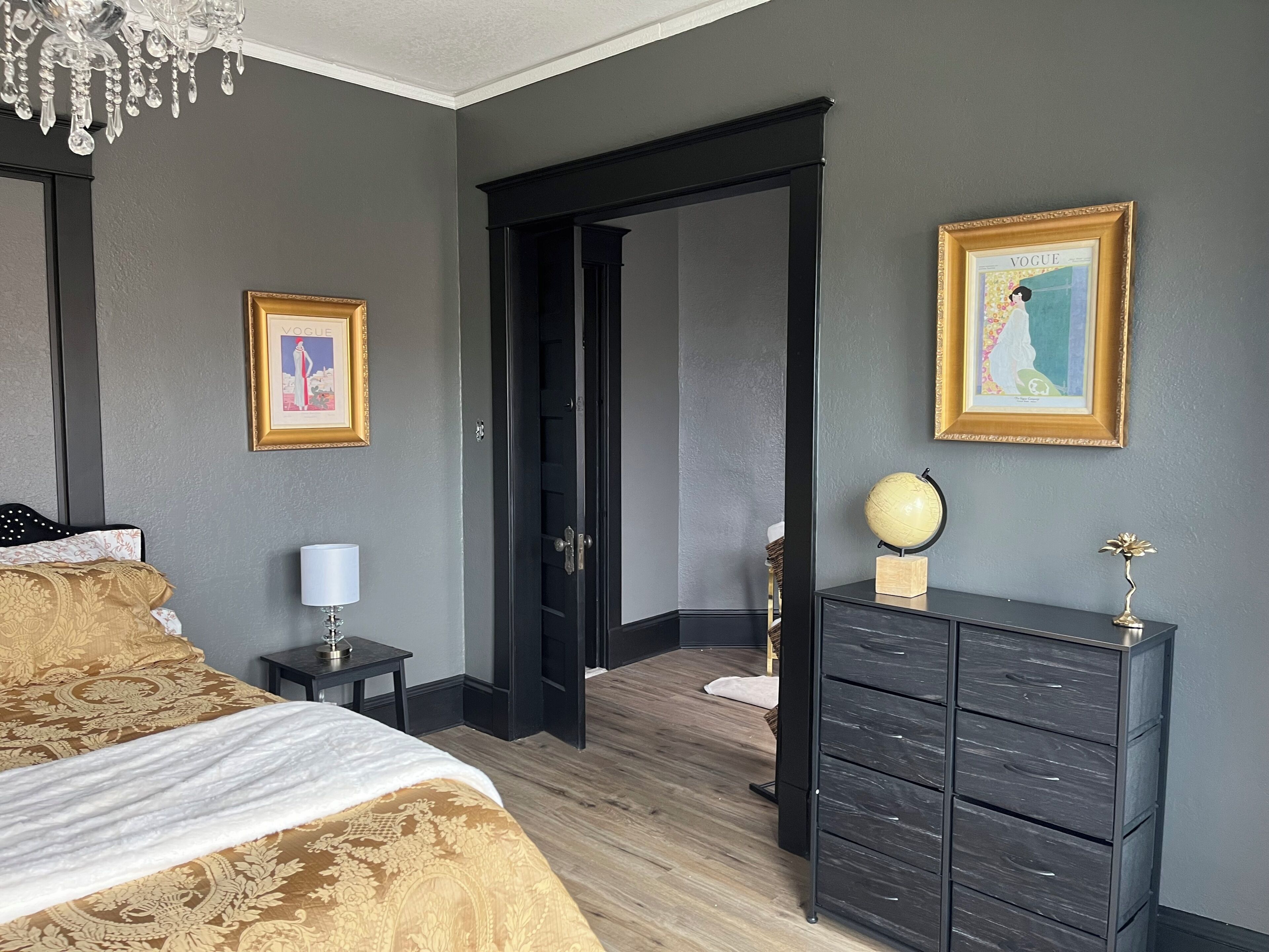 Une chambre à coucher comprenant un lit, une commode, une lampe, un tableau encadré et une porte donnant sur une autre pièce.