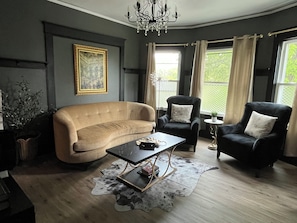Un salon avec un canapé beige, deux fauteuils, une table basse, un lustre et une grande fenêtre avec des rideaux.