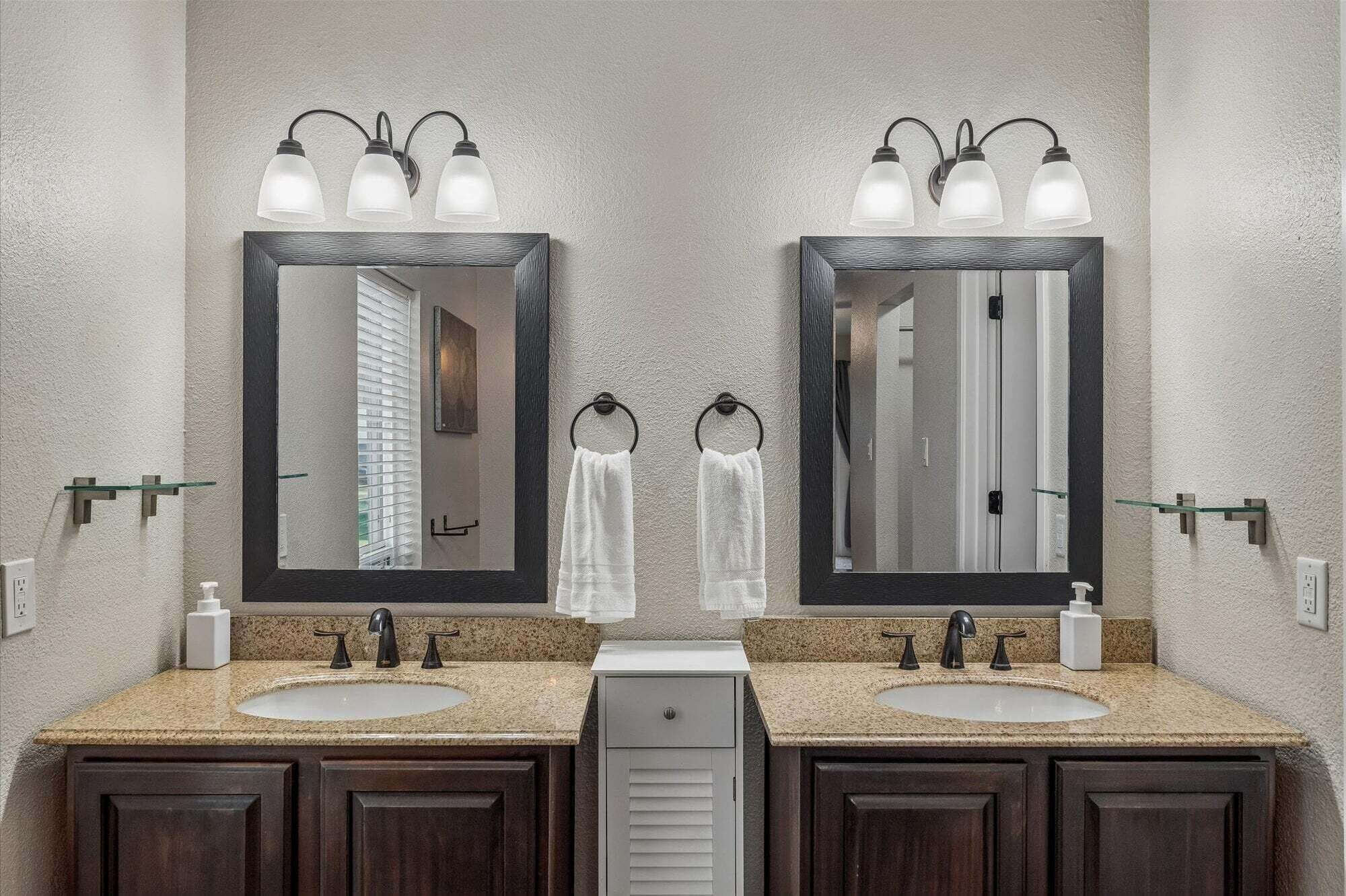 A bathroom with two sinks, a mirror, towel rings, and a frosted light fixture.