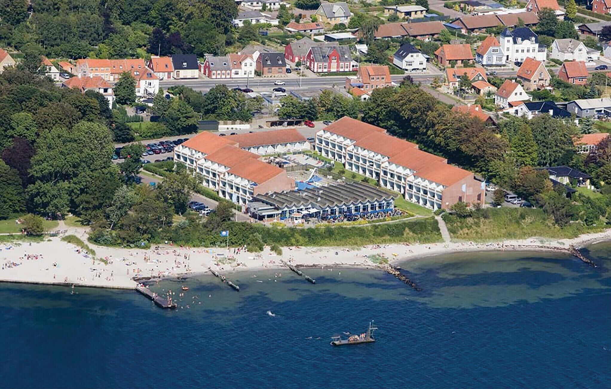 Vue aérienne d’un hôtel situé sur la côte, avec un bâtiment central, une plage et une étendue d’eau.