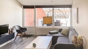 Een moderne woonkamer met een bank, een salontafel, een televisie en een groot raam met uitzicht op gebouwen.