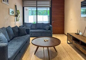A modern living room with a grey sofa, a wooden coffee table, and a wooden side table with a small screen. - Beautiful home in the jungle (Tulum)