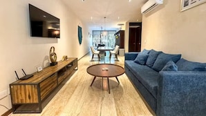 A modern living room with a flat-screen TV, a blue sofa, a wooden coffee table, and a dining area with chairs and a chandelier. - Beautiful home in the jungle (Tulum)
