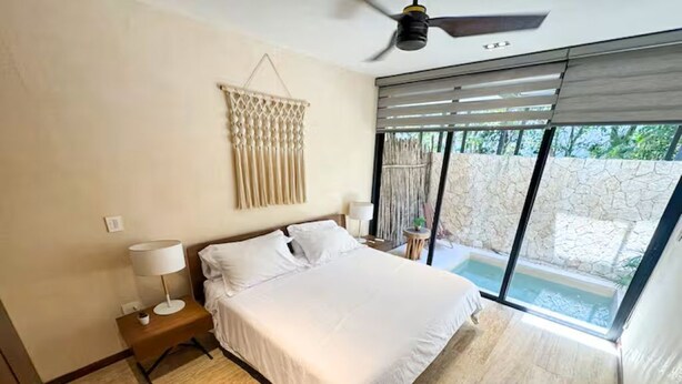 A bedroom with a bed, a nightstand, a lamp, a macrame wall hanging, and a view of a pool. - Beautiful home in the jungle (Tulum)