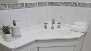 Un baño en esquina con un lavabo blanco, dos llaves, una toalla y una planta.
