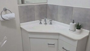 Un baño con un lavamanos blanco, dos grifos, un espejo sobre el lavamanos, un tozalengua y una pequeña planta en maceta.