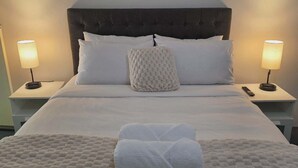 Una cama bien hecha con sábanas blancas, un suave plaid blanco y una toalla blanca doblada. Hay dos lámparas de noche a cada lado de la cama y tres cuadros abstractos encima.
