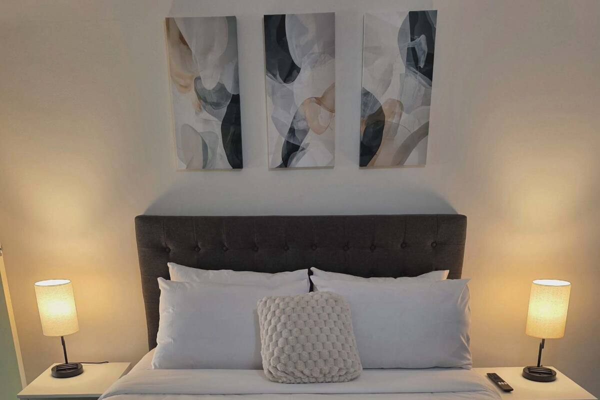 A neatly made bed with white linens, a fluffy white throw blanket, and a folded white towel. There are two bedside lamps on either side of the bed, and three abstract paintings above it.