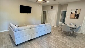 Ein Wohnzimmer mit grauem Sofa, einem Esstisch mit Stühlen, einem Deckenventilator, Einbauleuchten und einem Wand-Fernseher.