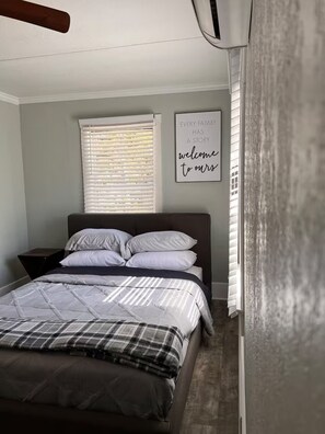 Ein Schlafzimmer mit Bett, einem Fenster mit Jalousien, einer Wand-Klimaanlage und einem gerahmten Schild.