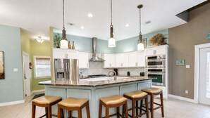 Una cucina moderna con mobili bianchi, un'isola centrale con sgabelli da bar, elettrodomestici in acciaio inossidabile e illuminazione a sospensione.