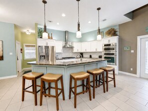 A modern kitchen with white cabinets, a central island with bar stools, stainless steel appliances, and pendant lighting. - Coastal Cotter | Sunset Patios + Pet Friendly! (Port Aransas)