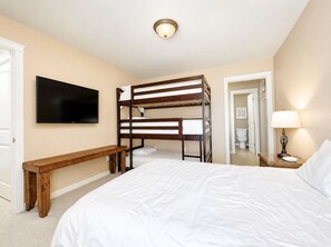 A bedroom with bunk beds, a flat-screen TV, a bedside lamp, and a wooden bench. - Coastal Cotter | Sunset Patios + Pet Friendly! (Port Aransas)