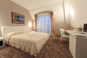 A hotel room with a bed, a desk, a chair, a painting on the wall, and a window with curtains.