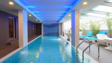 Una piscina con revestimiento azul, rodeada por un área de descanso con mobiliario blanco y techo de cristal.