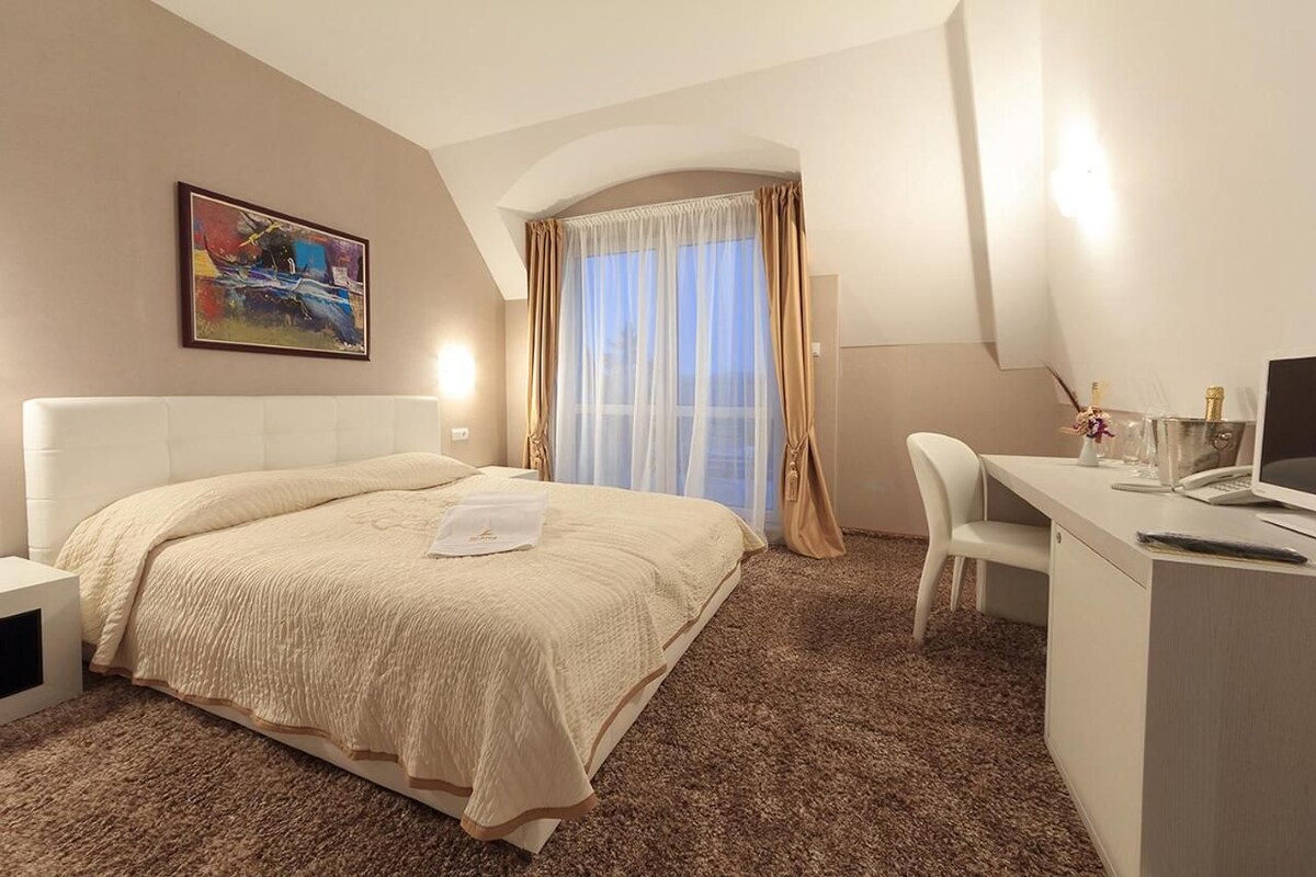 A hotel room with a bed, a desk, a chair, a painting on the wall, and a window with curtains.