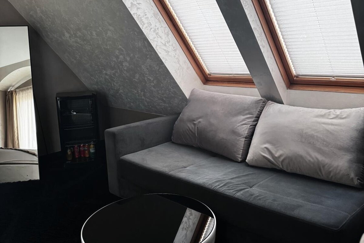 A modern loft bedroom with a sloped ceiling, a grey sofa, a round coffee table, and a built-in shelving unit.