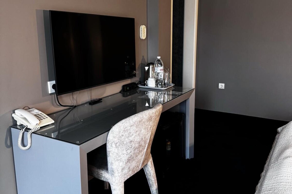 A hotel room with a desk, a flat-screen TV, a telephone, a chair, and a bed.